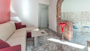 A modern living room with a beige sofa, a small wooden table, and a red floor lamp. There is a stone archway leading to a dining area with blue chairs.