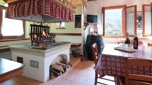 A rustic kitchen with a wood-burning stove, a dining table with chairs, and a view of the outdoors through large windows. - Holiday Home in Cison With Mountain Views (Cison di Valmarino)