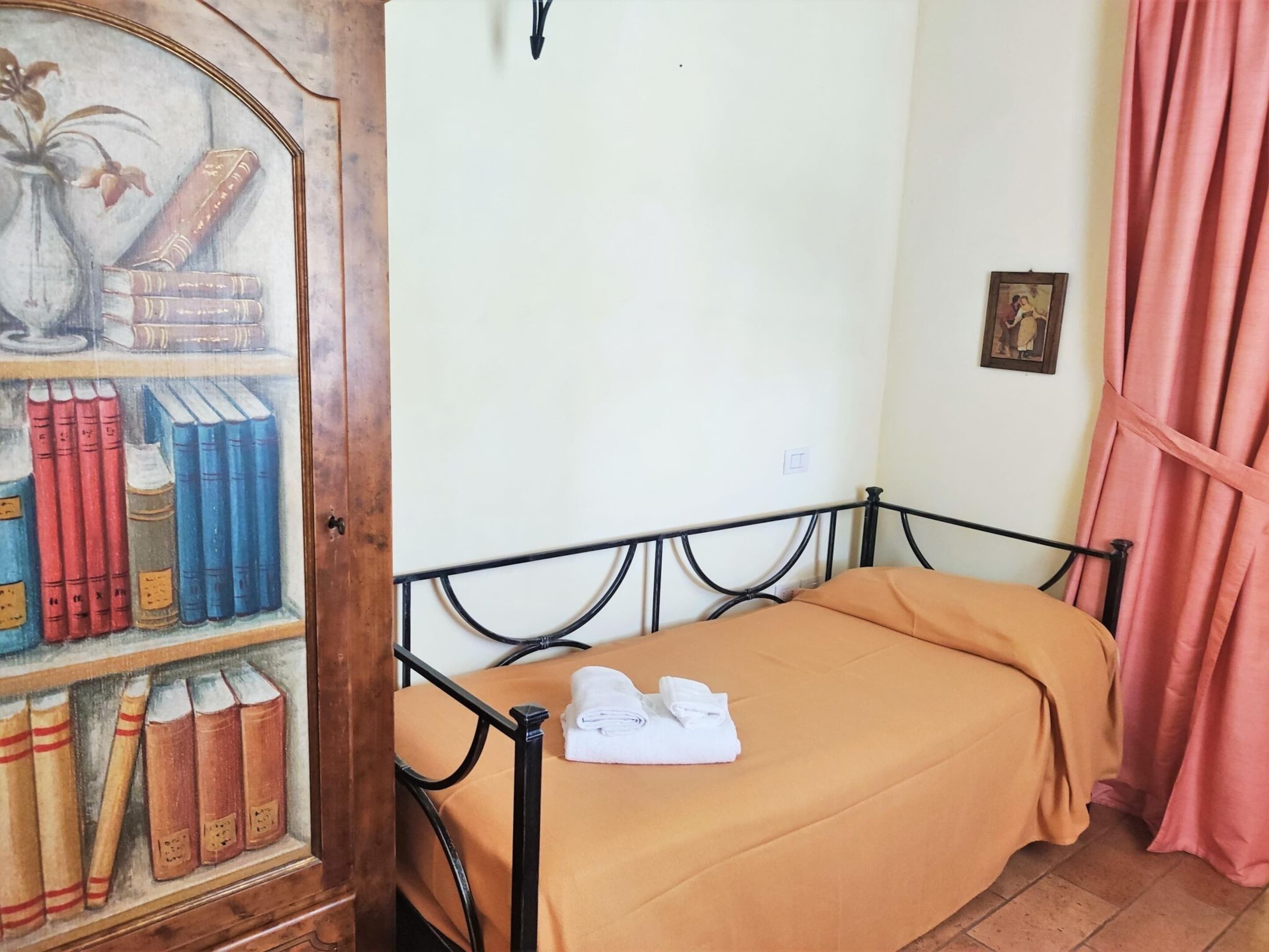 A single bed with a brown bedspread, a metal headboard, and a shelf with books and a vase of flowers.