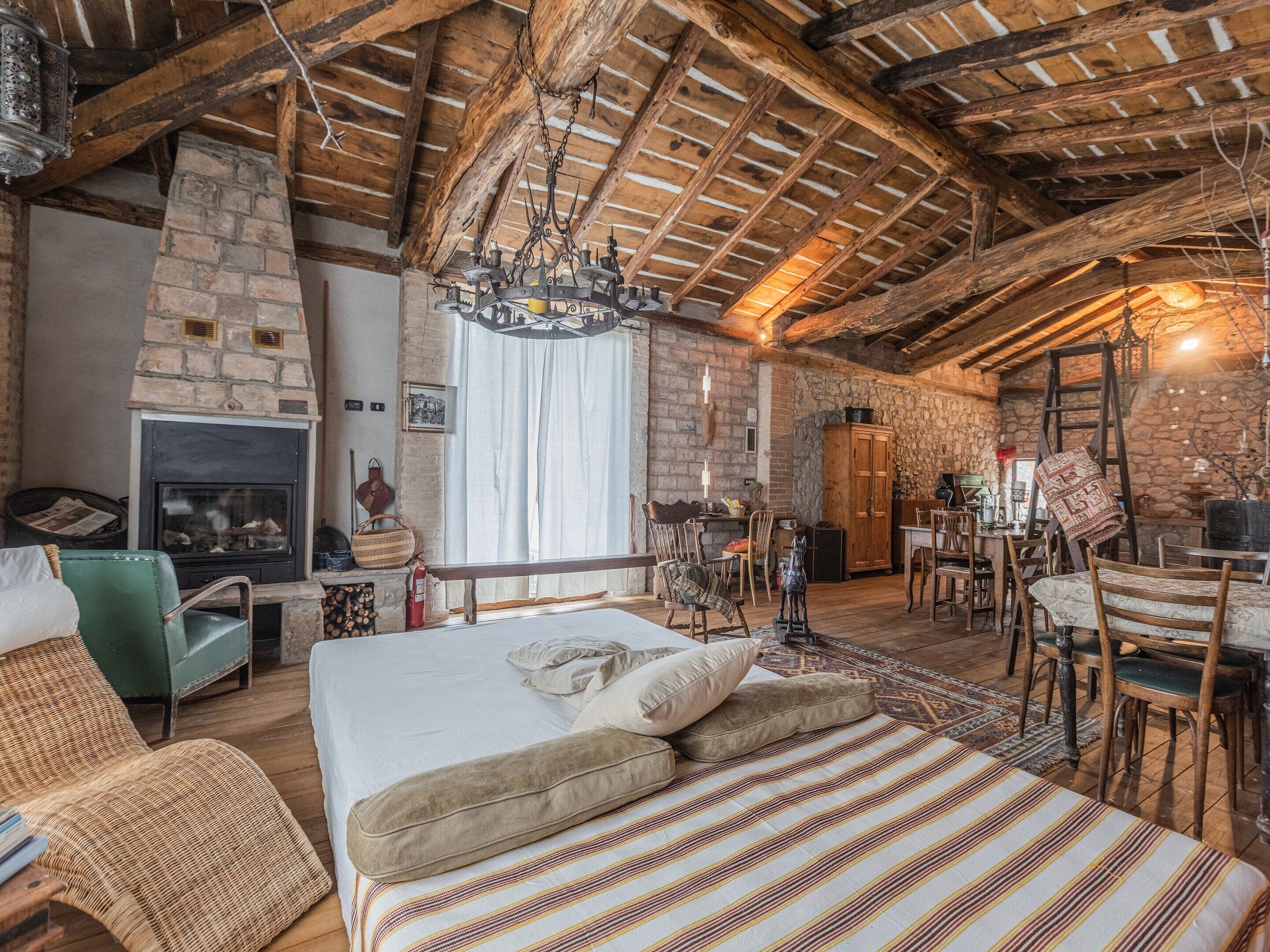 A rustic room with a stone fireplace, wooden beams, a chandelier, and a dining area.
