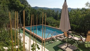 Piscina rectangular con agua azul cristalina, rodeada por una cerca de madera y una zona de césped artificial verde. Se encuentra una sombrilla blanca y una mesa con sillas cerca.