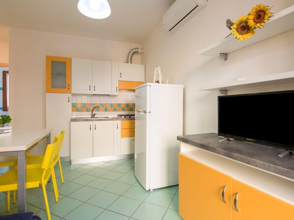 Een compacte keuken met witte en gele kastjes, een witte koelkast, een zwarte televisie op een grijze standaard, en een eethoek met een gele stoel.