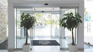 A modern entrance with glass doors, two potted plants, and a doormat with a logo. - Marlita Beach Hotel Apartments (Paralimni)