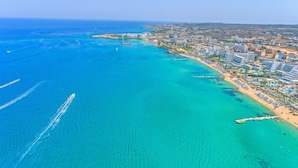 An aerial view of a coastal city with a clear blue sea, a sandy beach, and a densely built urban area with high-rise buildings. - Marlita Beach Hotel Apartments (Paralimni)