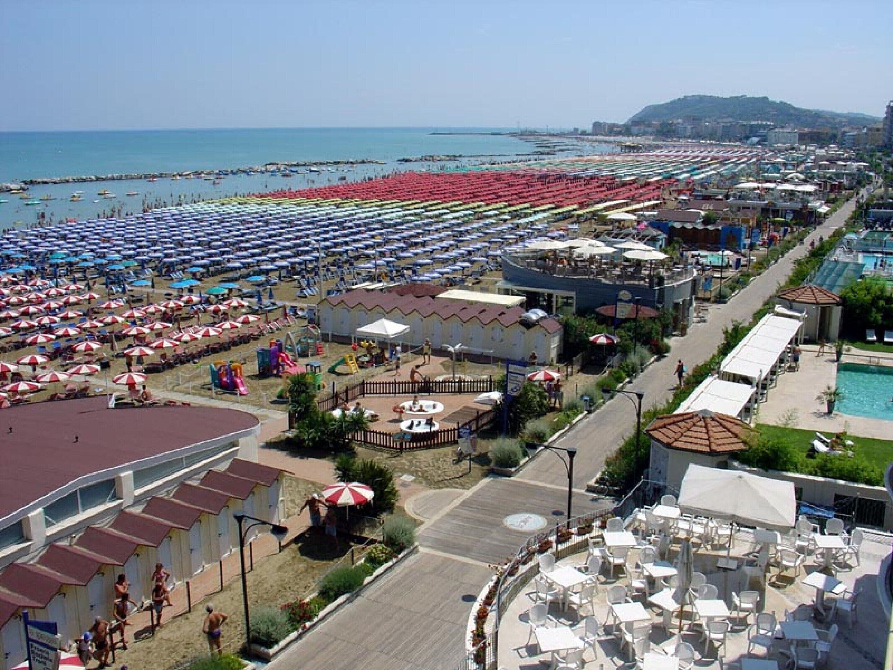 A coastal resort with numerous beach umbrellas, a swimming pool, and a variety of dining areas.