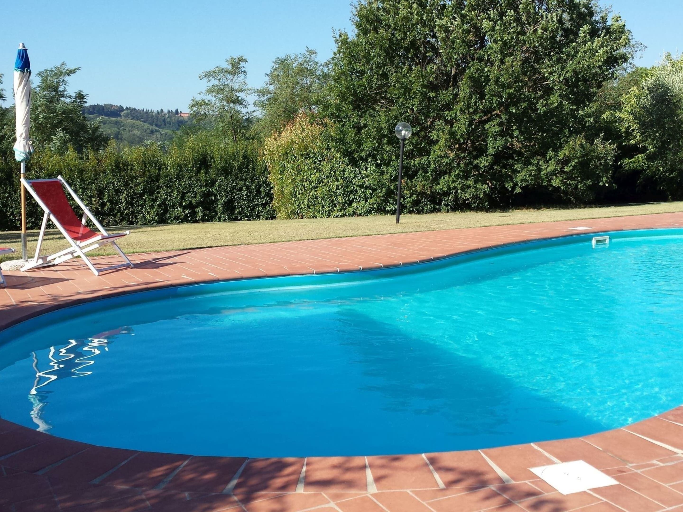 Piscina con una silla y un paraguas rojos, rodeada de un patio de ladrillo y abundante vegetación.
