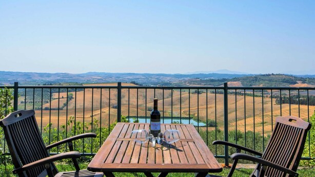 A wooden table with a bottle of wine and a glass, set against a scenic view of rolling hills and a lake, with metal chairs and a metal railing. - Apartment in Castelfiorentino With Pool (Castelfiorentino)