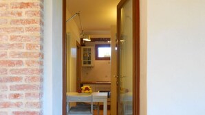 Un pasillo estrecho con una mesa y sillas blancas, una pequeña zona de cocina con ventana y una pared de ladrillo.