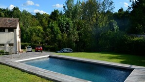 Una piscina rectangular con plataforma de madera, rodeada de césped y árboles.