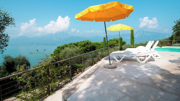 A poolside area with two yellow umbrellas, white lounge chairs, and a view of the sea and mountains. - Belvilla by OYO Gianni A Due Piao Due (Torri del Benaco)