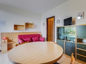 A room with a round wooden table, a pink sofa, wooden shelves, a television, and a cabinet with glass doors.