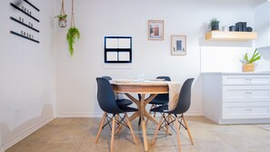 A modern kitchen with white cabinets, a wooden dining table, and black chairs. There are hanging plants, framed pictures on the wall, and a window with a view. - Designer Oasis: King Bed | Tranquil Backyard (Corpus Christi)