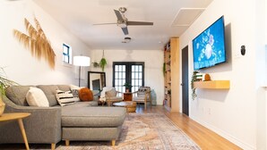 A modern living room with a grey sectional sofa, a ceiling fan, a large TV, and a bookshelf. - Designer Oasis: King Bed | Tranquil Backyard (Corpus Christi)