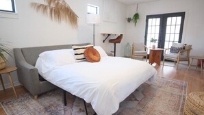 A bedroom with a bed, a grey sofa, a small table, and a chair. There is a window with a view of the outside. - Designer Oasis: King Bed | Tranquil Backyard (Corpus Christi)