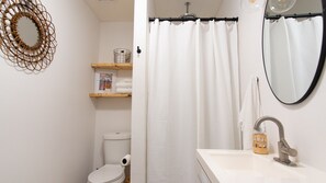 A bathroom with a toilet, a sink, a mirror, a shower curtain, and a shelf with towels. - Designer Oasis: King Bed | Tranquil Backyard (Corpus Christi)