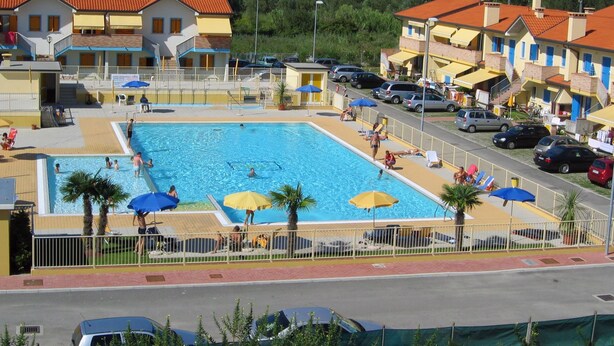 Una piscina rodeada de edificios con balcones, un área cercada y coches estacionados.