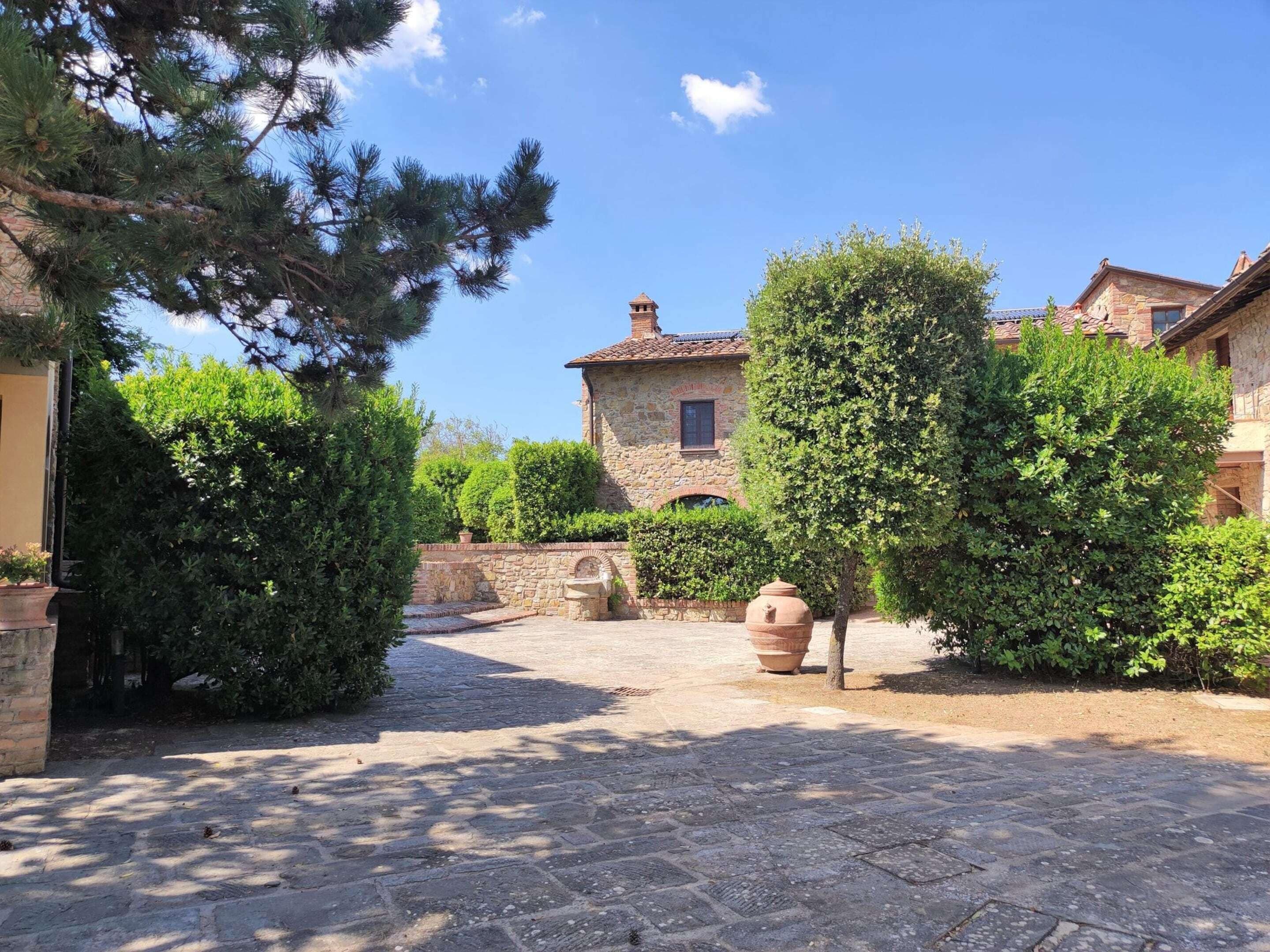 A courtyard with stone paving, a stone building with a tiled roof, and lush greenery.