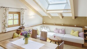 A cozy room with a high ceiling, wooden beams, a skylight, a sofa, a dining table, and a window with curtains.