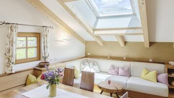 A cozy room with a high ceiling, wooden beams, a skylight, a sofa, a dining table, and a window with curtains.