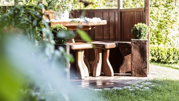 A wooden gazebo with a bench and table, surrounded by greenery and a mountainous backdrop.