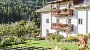 A two-story building with balconies adorned with flowers, surrounded by a well-maintained garden and a mountainous backdrop.