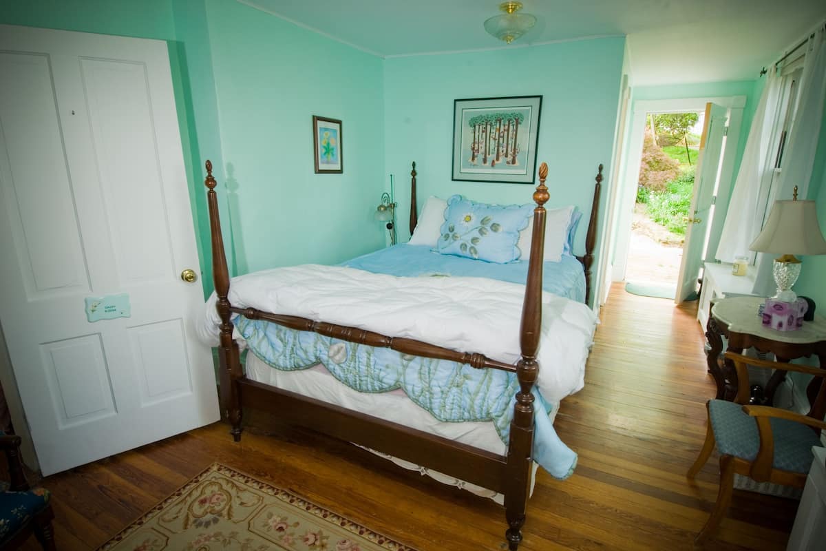 Quarto com cama de dossel, piso de madeira, porta com placa e pequena mesa com abajur.