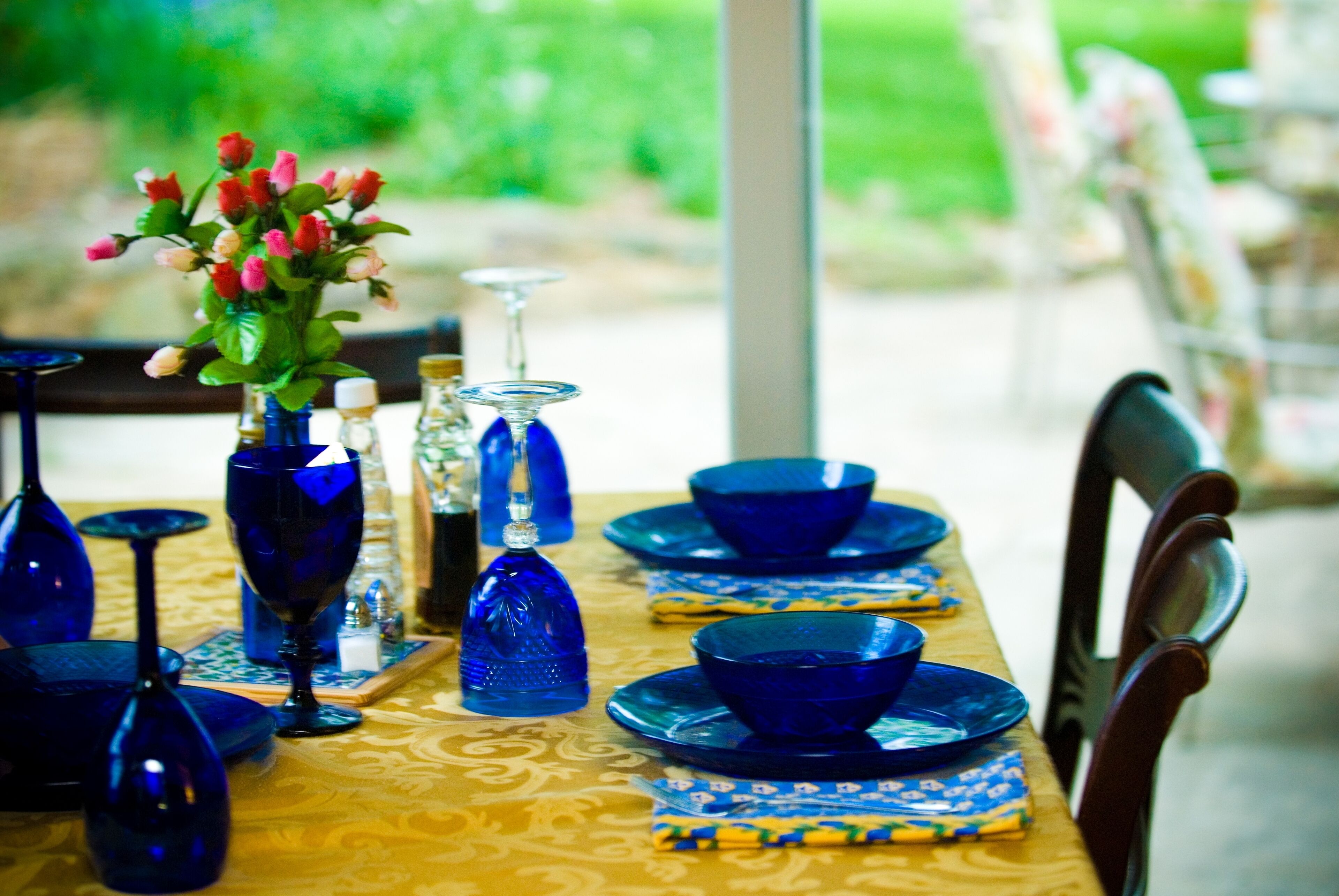 Ein Esstisch, gedeckt mit blauem Glas und Geschirr, einer Blumenvase und einer Tischdecke mit Muster.