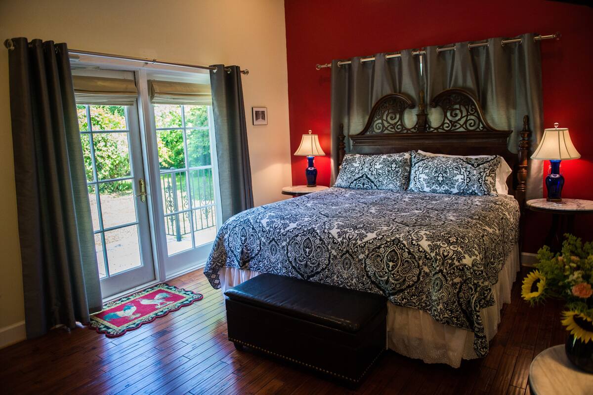 Quarto com cama grande, duas mesinhas de cabeceira com luminárias, um banco e vista através de uma porta de vidro.