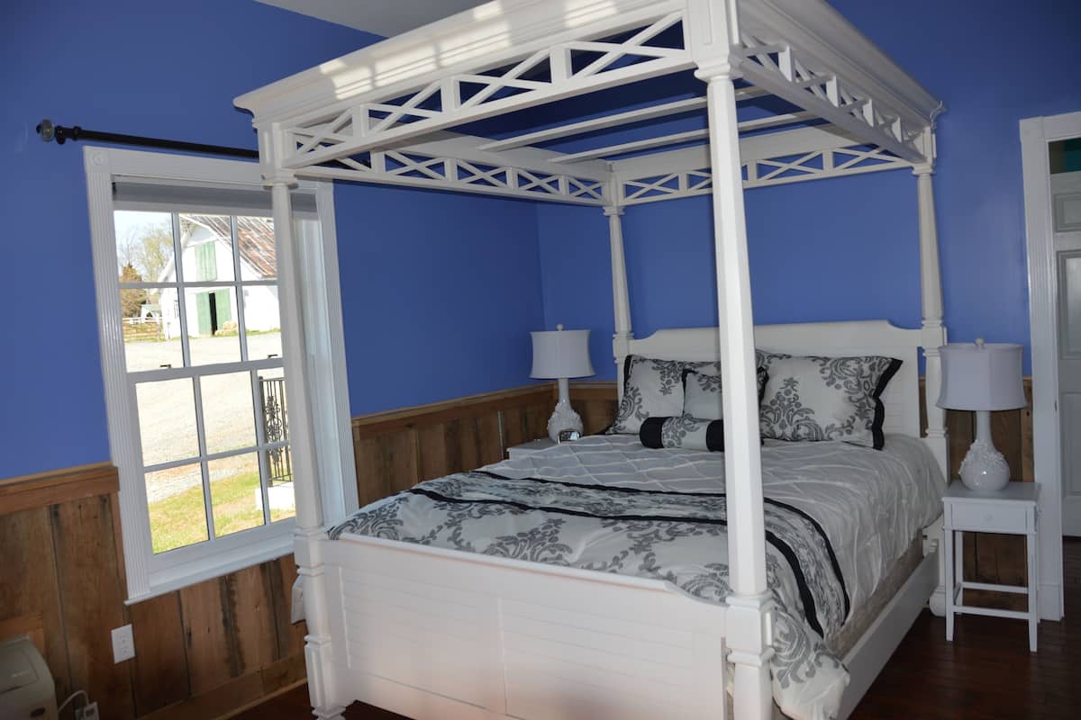 Uma cama de dossel com dossel, roupa de cama branca, duas mesas de cabeceira com abajures e uma janela com vista para o exterior.
