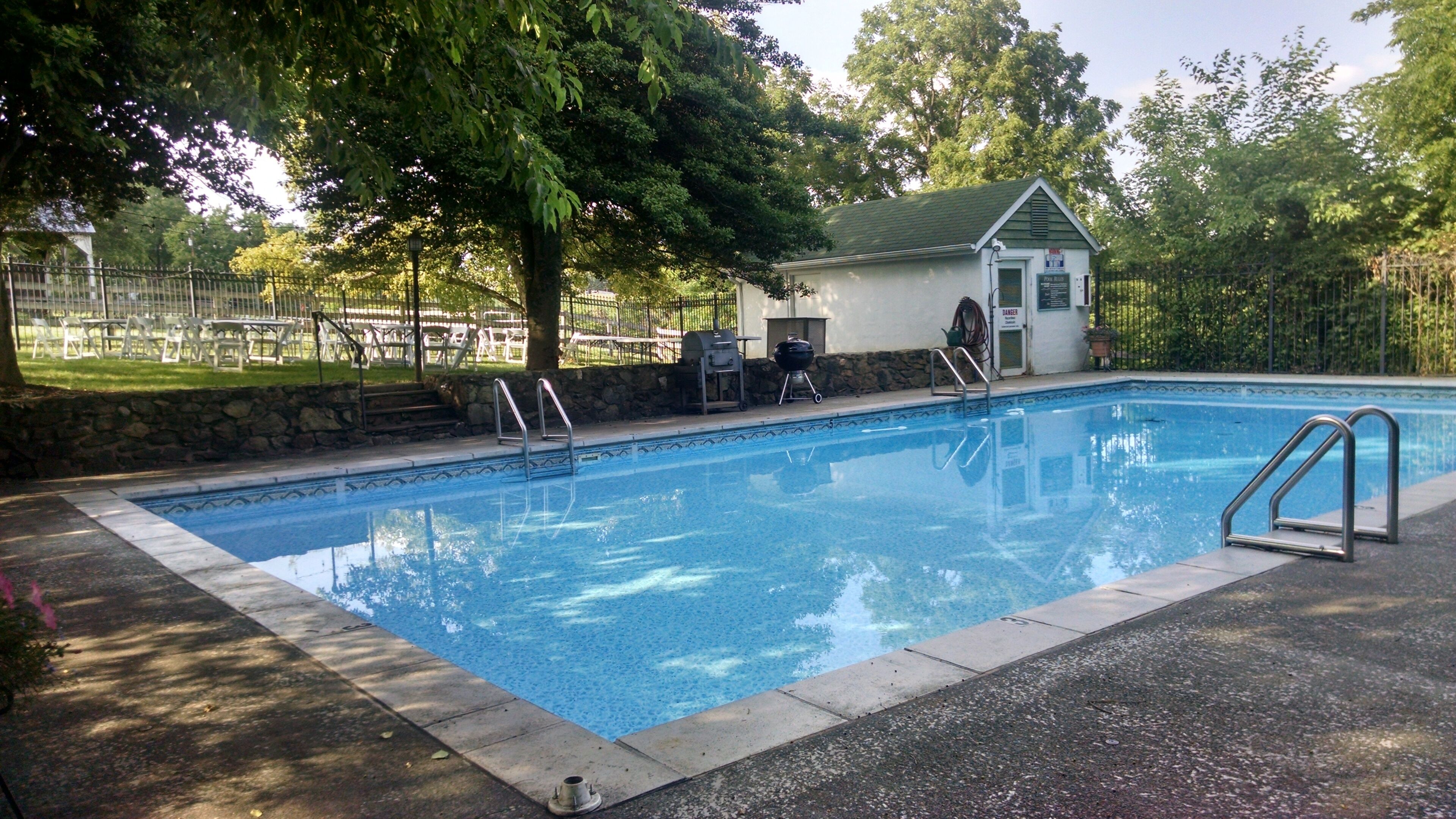 A rectangular swimming pool with a metal ladder on one side, surrounded by a stone wall and a grassy area with chairs and tables.