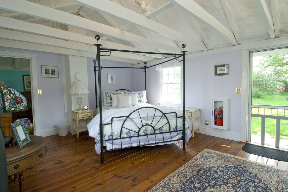 Uma cama com dossel em um quarto com piso de madeira, tapete estampado e vista para o verde lá fora.