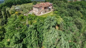 Ein Steinhaus mit Ziegeldach, umgeben von üppiger Vegetation und Hügeln im Hintergrund.