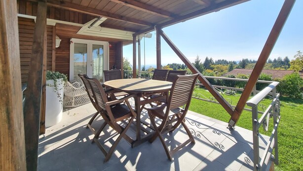 A covered patio with a wooden table and chairs, a white railing, and a view of greenery and buildings in the distance. - Chalet in Marliana Near Center (Marliana)