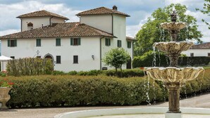 A fountain with water cascading down its tiers, surrounded by a circular stone basin. In the background, there is a white building with a tiled roof and green shutters, set against a backdrop of lush greenery and a clear sky. - Belvilla by OYO Villa Terra (Reggello)