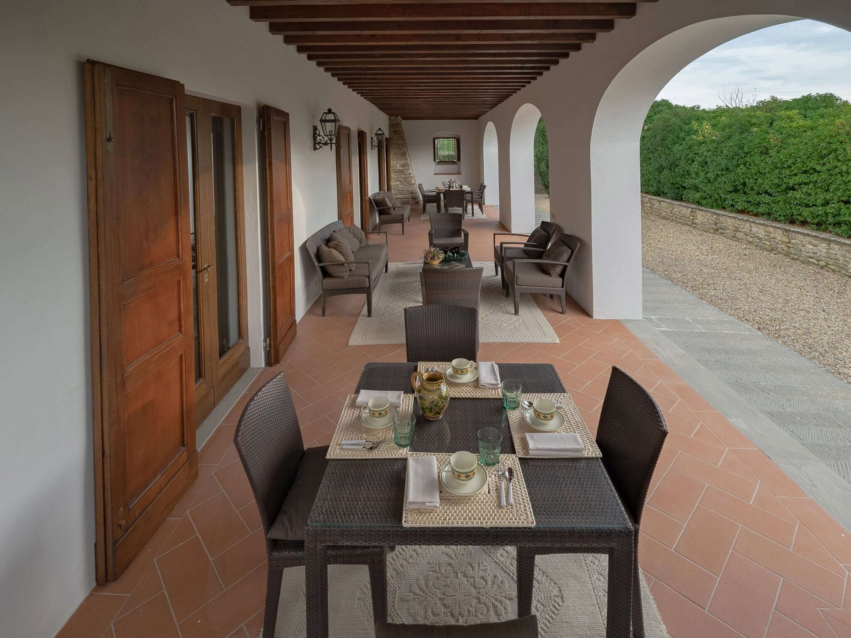 Un espacio de comedor al aire libre con techo, amueblado con ratán y suelo de piedra, que ofrece vistas a zonas verdes.
