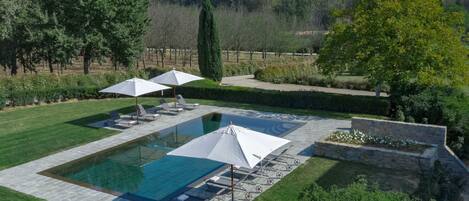A rectangular swimming pool with a stone border, surrounded by a well-manicured lawn and a stone wall. There are lounge chairs and umbrellas under a pergola.