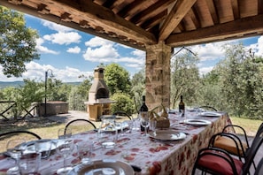 Un comedor al aire libre con una mesa preparada para una comida, un horno de ladrillo y vistas del paisaje circundante.