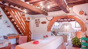Una habitación rústica con una escalera de madera, una mesa con un cuenco rojo y vistas a un muro de piedra a través de un arco.