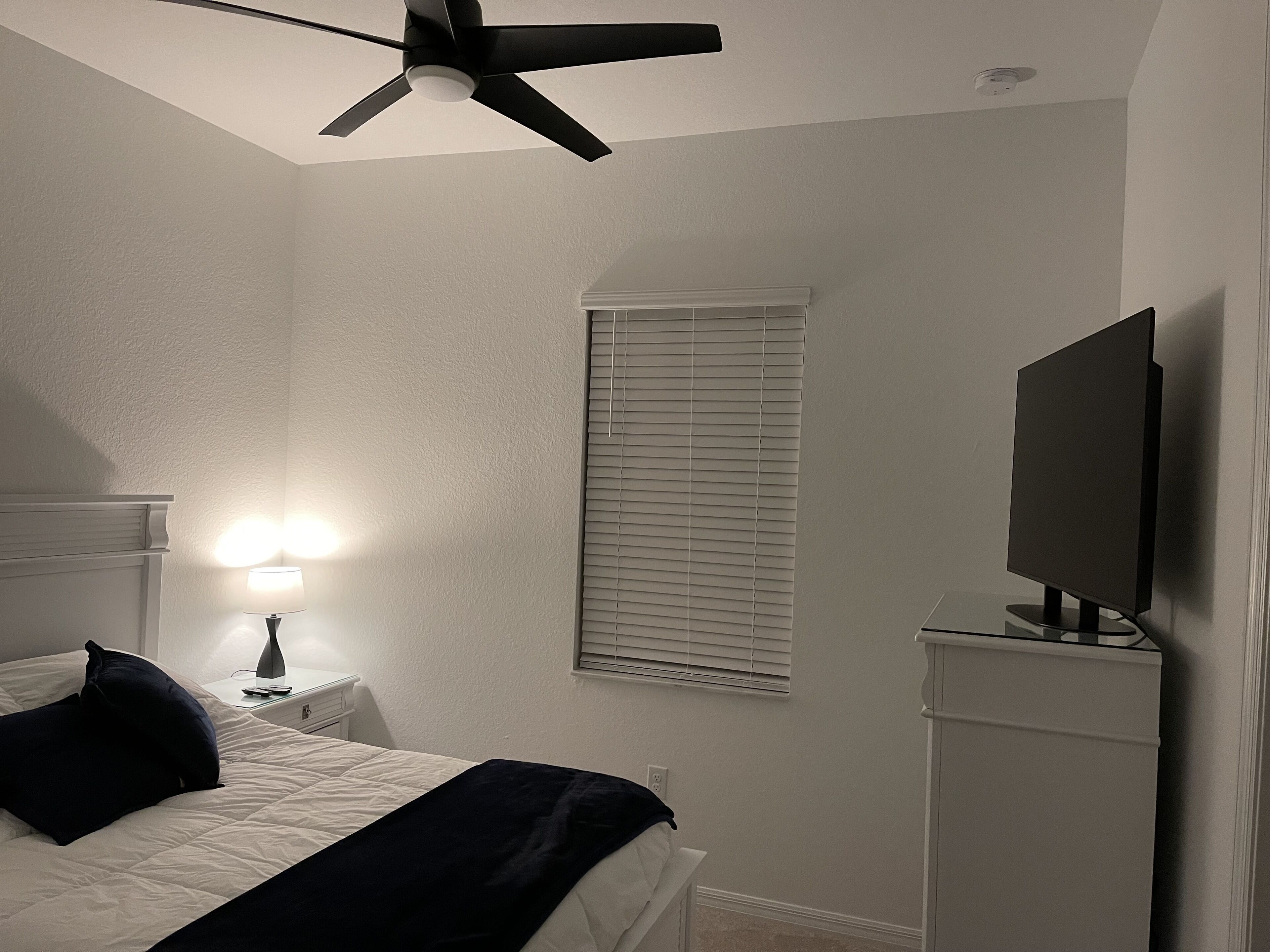 Une chambre à coucher avec un lit, un ventilateur de plafond, une lampe de chevet, un téléviseur posé sur un meuble et une fenêtre avec des stores.