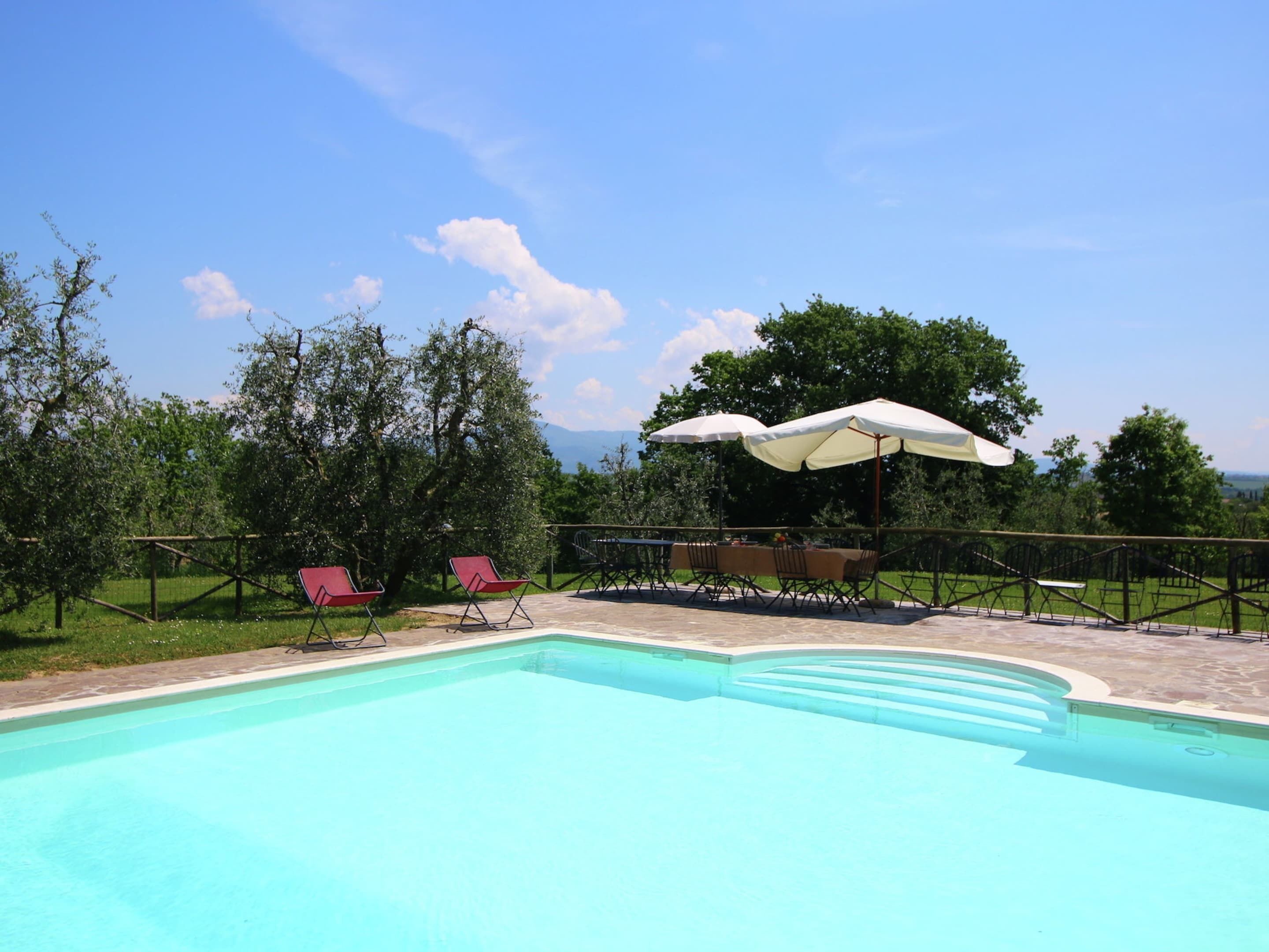 A swimming pool with red chairs and umbrellas, surrounded by trees and a view of the mountains.