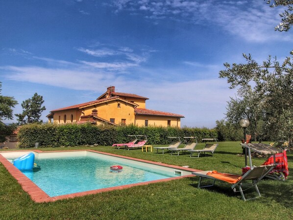 Zona de piscina con sillones de descanso, una piscina y un edificio grande al fondo.