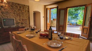 Un comedor con una mesa preparada para cuatro personas, una pared de piedra, un televisor y vistas al exterior con vegetación.