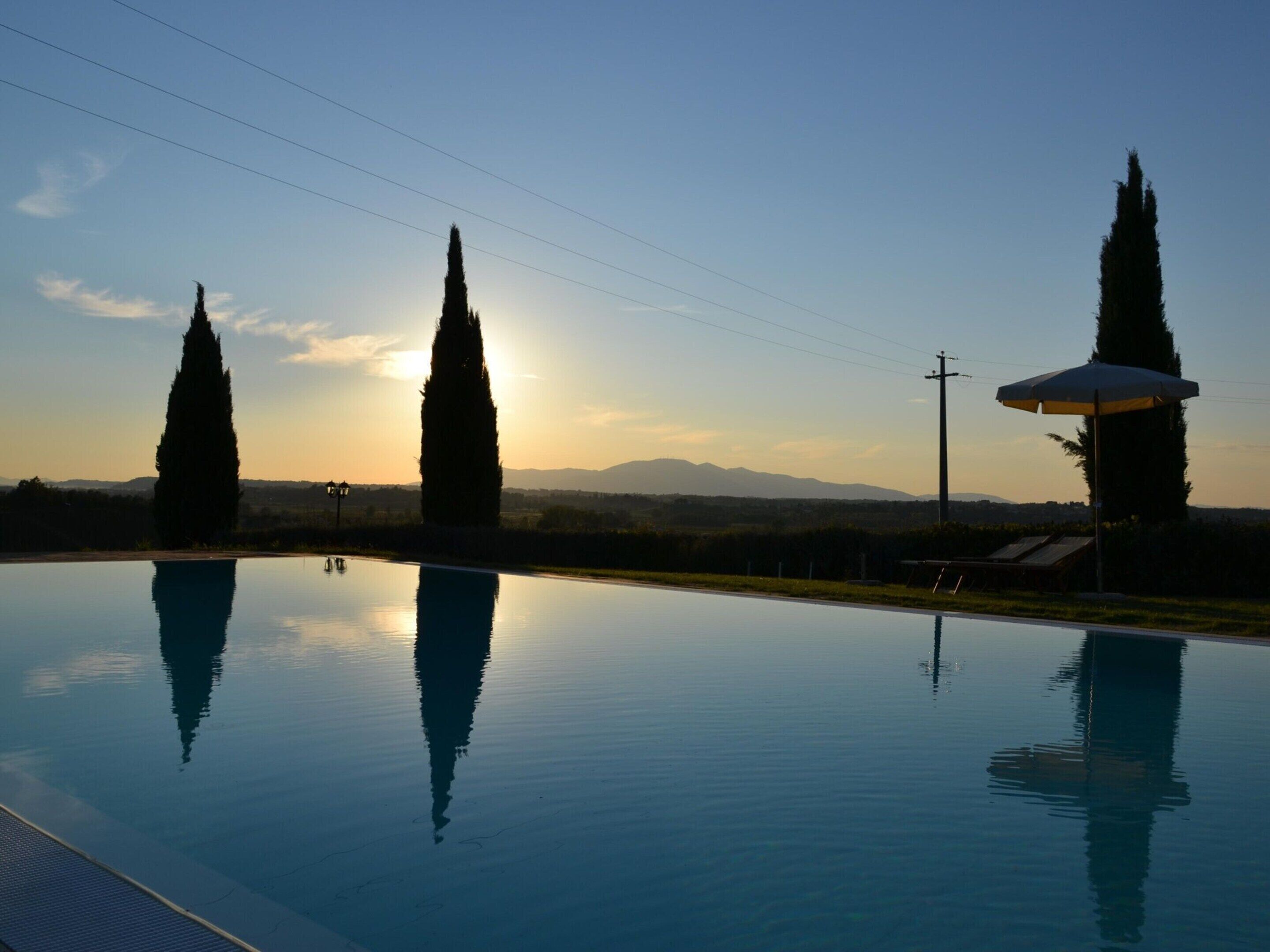 Ein ruhiger Pool mit hoch aufragenden Zypressen und einem Sonnenuntergang im Hintergrund.