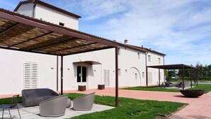 Een overdekt buitengebied met stoelen en een vuurkorf, grenzend aan een wit gebouw met een pannendak en een pergola.
