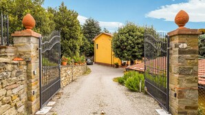 Una strada sterrata conduce a una casa gialla con un muro in pietra e cancelli in metallo.