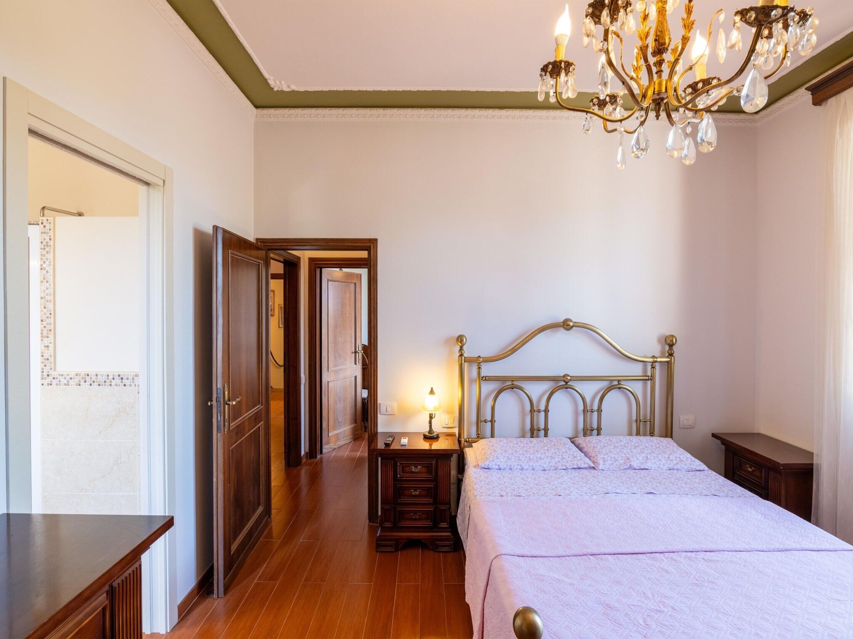 Een slaapkamer met een houten bed, een nachtkastje met een lamp, een kroonluchter en een deur die naar een andere kamer leidt.