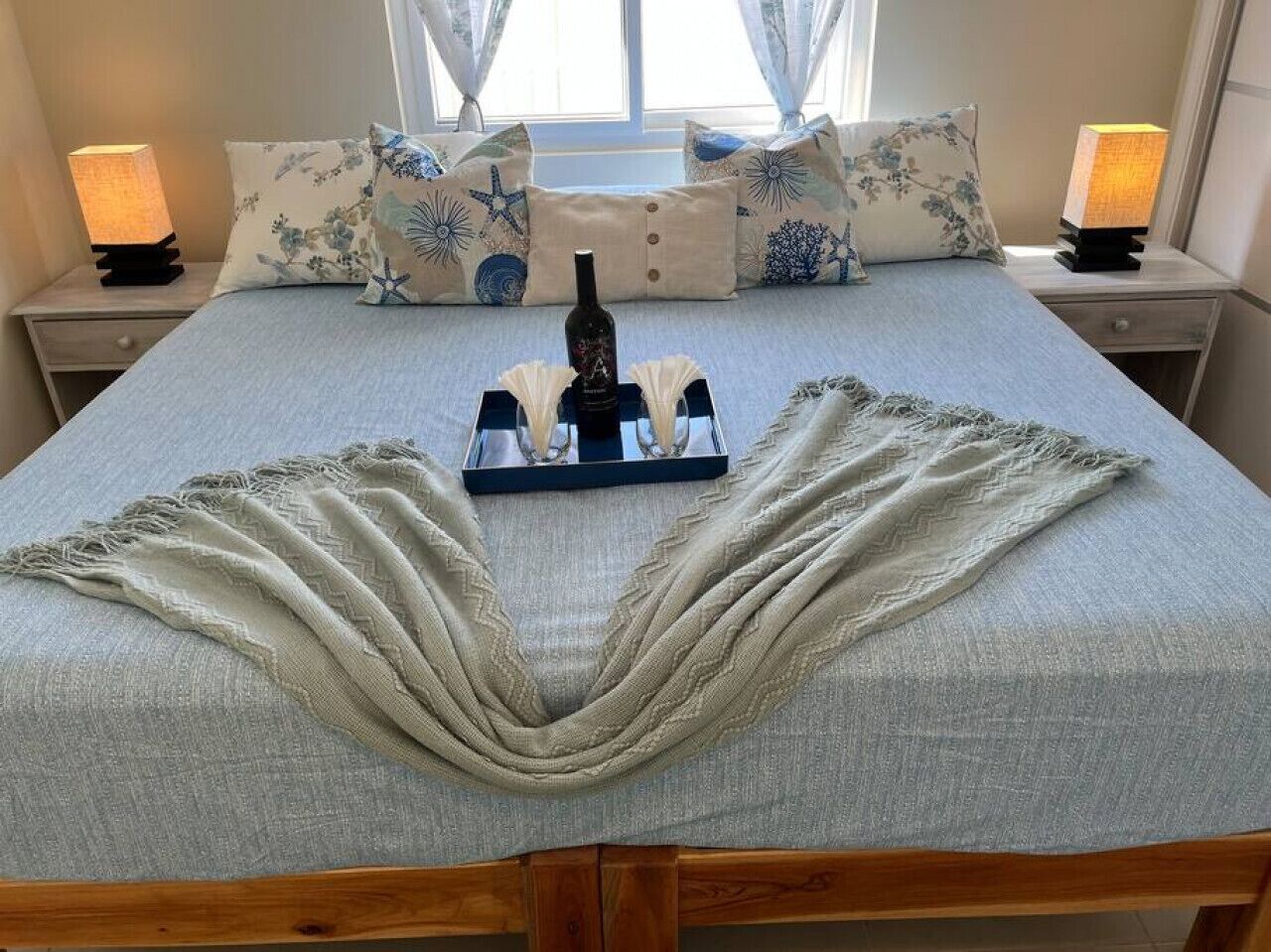 A neatly made bed with a decorative blanket, a bottle, and glasses on a tray, flanked by matching pillows and bedside lamps.