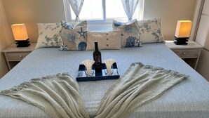 Een netjes opgemaakt bed met een decoratieve deken, een fles en glazen op een dienblad, omringd door bijpassende kussens en nachtlampjes.