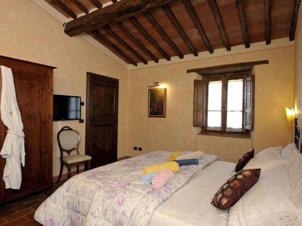 Une chambre avec un plafond en bois, un lit recouvert d’une couette à motifs floraux, une chaise, une armoire en bois, un téléviseur et une fenêtre avec des rideaux.