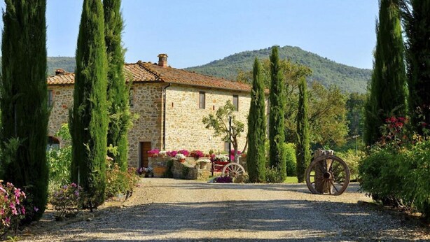 A stone building with a gravel driveway, surrounded by tall cypress trees and a mountain in the background.